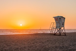 A beach at sunset, the sun is just above the horizon still. A lifeguard station stands to the right side of the image.