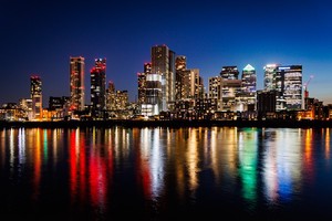 The tall buildings of London shot over the River Thames at night. The buildings arte lit with lights which reflect off the smooth surface of the river.