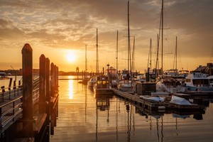 A photo of a harbour. On the left a pier stretches out into the image with post spaced periodically. To the right are a number of yachts docked. Between the two the sun rises into a cloudy sky. The light reflects off the clouds and the rippled water bathing the image in an orange glow.